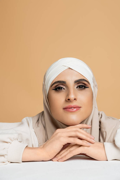 positive and stylish muslim woman with makeup sitting at white table in hijab and smiling at camera - Photo, Image