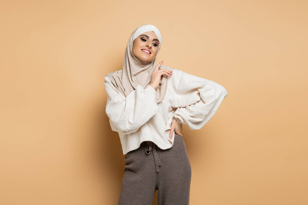 mujer musulmana alegre y moderna en hijab y atuendo casual de moda posando con la mano en la cadera en beige - Foto, Imagen