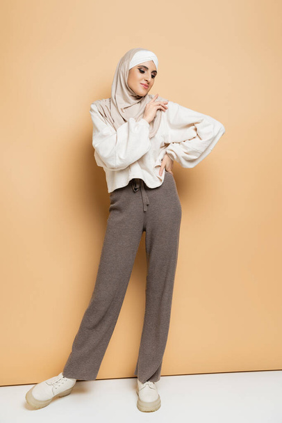 fashionable muslim woman in hijab, white sweatshirt and grey pants posing with hand on hip on beige - Photo, Image