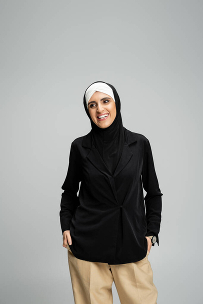 succesvolle moslim zakenvrouw in hijab en zwarte jas, met stralende glimlach op grijs - Foto, afbeelding