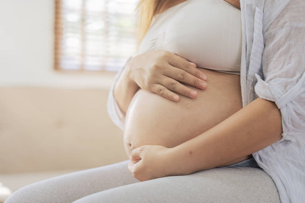 Onnellinen nainen raskaus istuu sohvalla ja pitää hänen vatsa olohuoneessa. - Valokuva, kuva