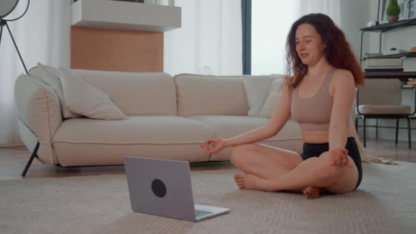 Evde dizüstü bilgisayarlı web kamerası yogası kullanarak bacak bacak bacak üstüne atmış bir kadın var. - Video, Çekim