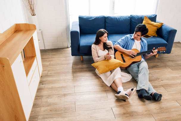 Attraktive neue Eheleute sitzen auf dem Boden und genießen es, im Wohnzimmer des neuen Heims zu singen und Gitarre zu spielen. Eine Familie verbringt nach dem Einzug in ein neues Zuhause viel Zeit miteinander. - Foto, Bild