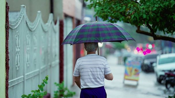 Az idősebb férfi elmegy otthonról esőben, kinyitja az esernyőt és becsukja maga mögött a kaput. A végzős az utcán sétál. - Fotó, kép