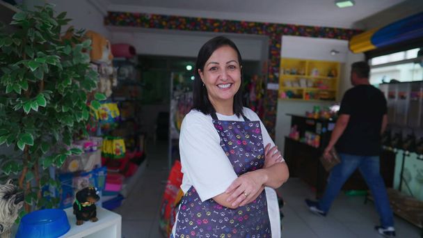 Happy woman entrepreneur of PetShop smiling while crossing arms confidently. Portrait of a small business owner wearing apron standing in front of Storefront - Photo, Image