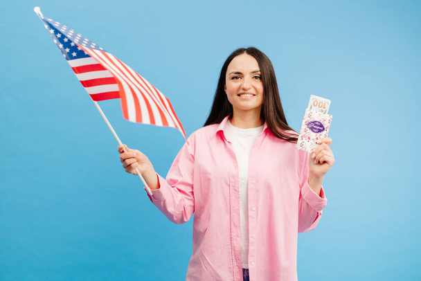 Νεαρή μελαχρινή με ροζ πουκάμισο ποζάρει σε μπλε φόντο στο στούντιο με αμερικάνικη σημαία και εκατό δολάρια μετρητά με διαβατήριο. Έννοια της επιτυχίας, τα ταξίδια, η γνώση της ξένης γλώσσας. - Φωτογραφία, εικόνα