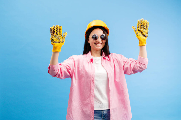 Γυναίκα σε κίτρινο κράνος και γάντια που δείχνουν και gesturing κατά μπλε φόντο στούντιο. - Φωτογραφία, εικόνα