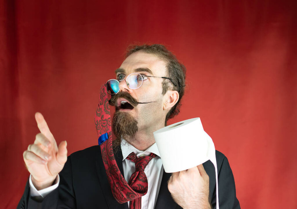Vtipný muž s brýlemi, červenou kravatou na hlavě a toaletním papírem v ruce vzhlíží s červeným pozadím - Fotografie, Obrázek