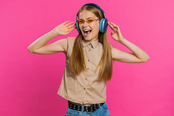 Happy woman in headphones listening music and dancing disco fooling around having fun expressive gesticulating hands relaxing on party. Blonde girl in sunglasses isolated on studio pink background - Photo, Image