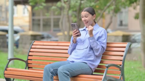 ベンチに屋外で座っている間,スマートフォンで成功を祝うヒスパニック女性 - 映像、動画