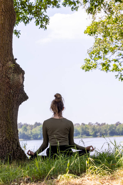 Спокойный и спокойный момент, запечатленный в природе, где молодая брюнетка изящно сидит в пышной зеленой траве рядом с величественным деревом. Она смотрит на мирное лесное озеро, ее глаза - Фото, изображение