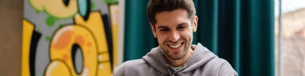Happy young brunette man holding mobile phone while sitting at the cafe table indoors - Foto, imagen