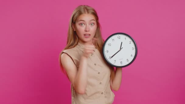 It is your time. Young attractive woman showing time on wall office clock, ok, thumb up, nice good, approve, start, pointing finger at camera. Blonde girl isolated on pink studio background, indoors - Footage, Video