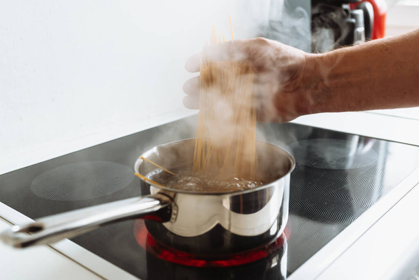 чоловік готує макарони спагеті вдома на кухні. чоловік готує в коричневому фартусі, пробує готовність спагеті, тримає спагеті на спеціальній ложці над киплячою водою. Італійська концепція домашньої кухні - Фото, зображення