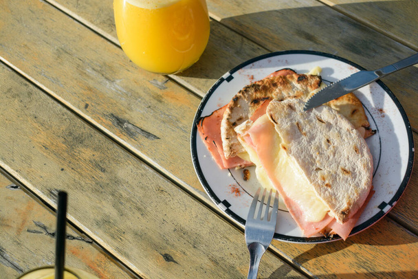 bovenaanzicht van ham en kaas sandwich met Arabisch brood geserveerd op bord met sinaasappelsap op houten tafel buiten bij zonsondergang, eten en drinken concept, kopieer ruimte. - Foto, afbeelding