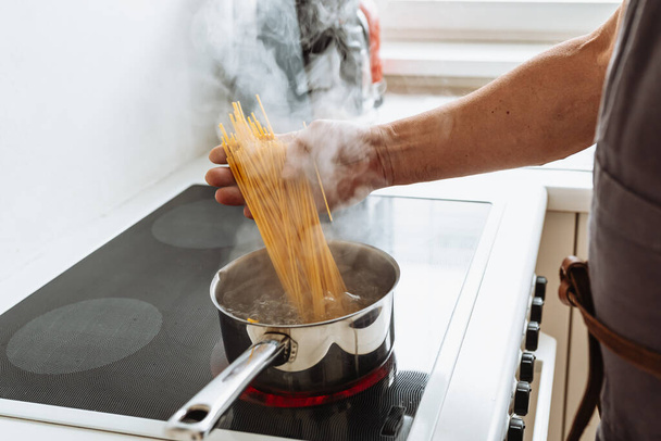 mans main trempettes spaghetti dans l'eau bouillante dans une casserole debout sur plaque vitrocéramique, dans la cuisine de la maison - Photo, image