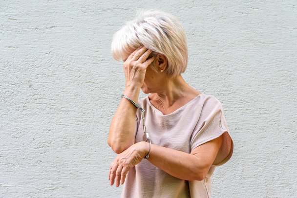 Літня жінка 60-65 років ховає обличчя, наручники на руках, сірий фон.  - Фото, зображення