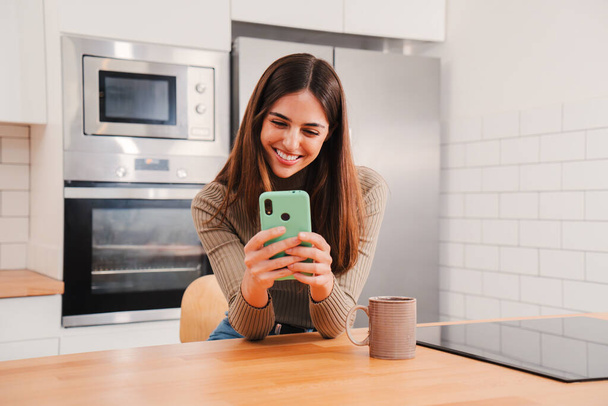 Happy young Caucasian woman using her mobile phone smiling at screen, taking a break sitting at home kitchen. Portrait of joyful lady chatting on cell phone or getting good messages on social media - Photo, Image