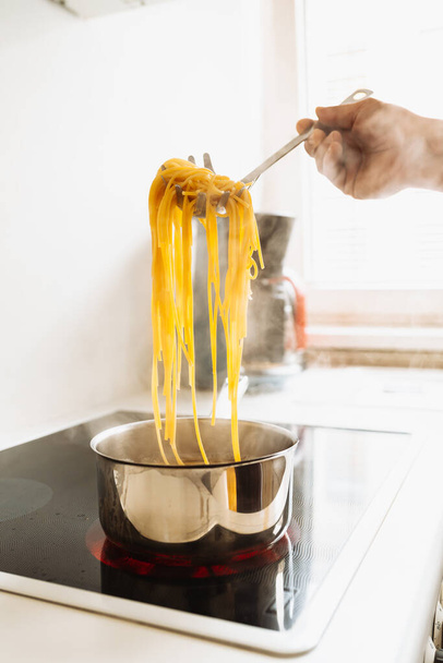 mężczyzna gotuje makaron spaghetti w domu w kuchni. mężczyzna gotuje w brązowym fartuchu, próbuje spaghetti gotowość, trzyma spaghetti na specjalnej łyżce nad wrzącą wodą. Koncepcja włoskiej kuchni domowej - Zdjęcie, obraz