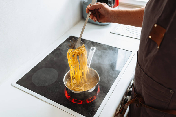Adam evde, mutfakta makarna pişiriyor. Kahverengi önlüklü bir adam spagettiyi dener, spagettiyi kaynamış suyun üzerinde özel kaşıkta tutar. İtalyan ev yemeği konsepti - Fotoğraf, Görsel