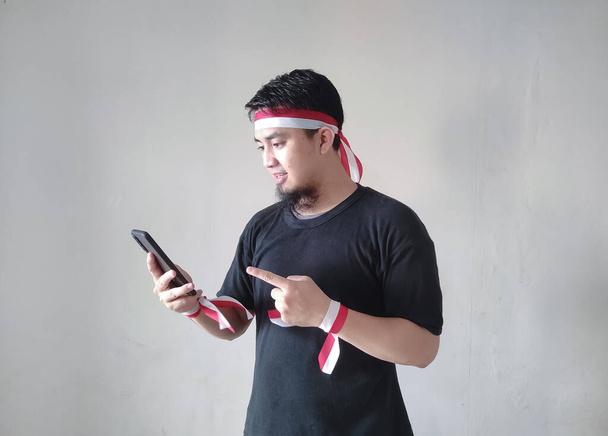A young Asian man with happy successful expression while holding his mobile phone and wearing black t-shirt, flag headband. Indonesia independence day concept. - Photo, Image