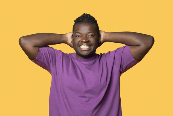 リラックスしたポーズに頭の後ろに立ち,黄色い背景の上に立っているカメラで笑い,紫色のTシャツを着ているトゥーシーなアフリカ系アメリカ人の男の肖像. スタジオショット - 写真・画像