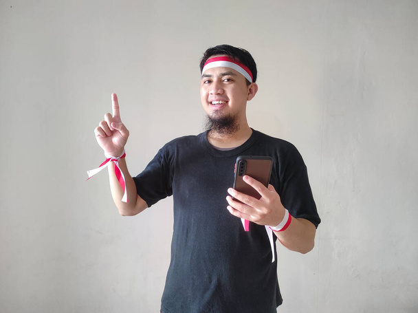 Молодий азіатський чоловік з щасливим успішним виразом тримає свій мобільний телефон і носить чорну футболку, флагманську пов'язку. Концепція Дня незалежності Індонезії. - Фото, зображення
