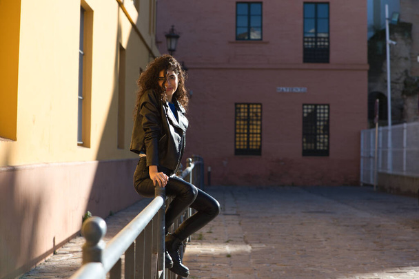 Jonge en mooie Spaanse brunette vrouw met krullend haar en zwarte leren kleding zit op een reling in de straat. De vrouw is gelukkig en glimlachend. - Foto, afbeelding
