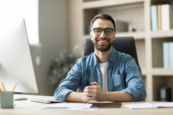 Sukces przedsiębiorcy. Portret szczęśliwego tysiącletniego mężczyzny pozującego w biurze, przystojnego, uśmiechniętego młodego biznesmena siedzącego przy biurku z komputerem i patrzącego w kamerę, kopiującego przestrzeń - Zdjęcie, obraz