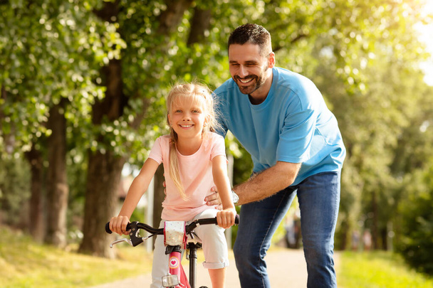 Loving european middle aged father and his pretty daughter spending time together oudoors, dad teaching child how to ride a bicycle, walking in park - Photo, Image