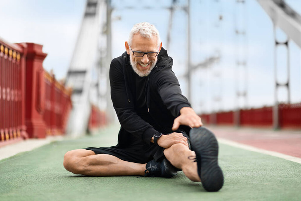 Šťastný pohledný šedovlasý, dobře padnoucí starší sportovec v černém sportovním oblečení, sedící na pozemním mostě, dosahující rukou k nohám, muž v důchodu protahuje tělo, zatímco ráno cvičí venku - Fotografie, Obrázek