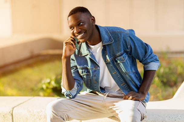 Gut aussehender junger schwarzer Kerl in lässigem Outfit sitzt auf der Straße, telefoniert und lächelt, schaut auf den Kopierraum. Glücklicher Mann Student wartet auf seine Freunde, telefonieren - Foto, Bild