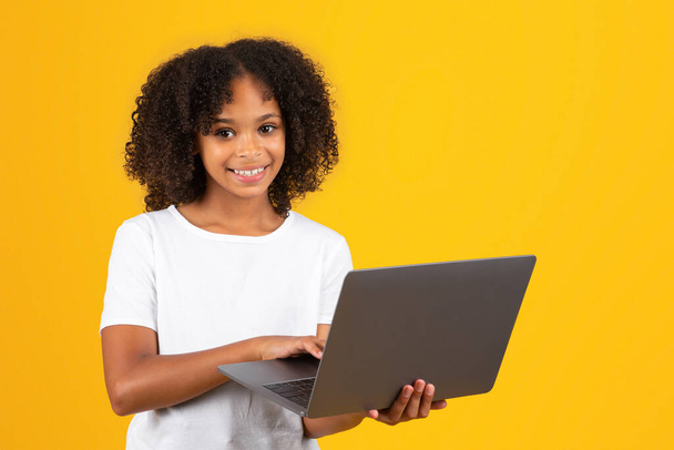 Положительный подросток черного зрачка девочка в белой футболке использует ноутбук, вач видео урок, изолированные на желтом фоне, студия. Обучение в школе дистанционно, объявление и предложение, гаджет для образования, образ жизни - Фото, изображение