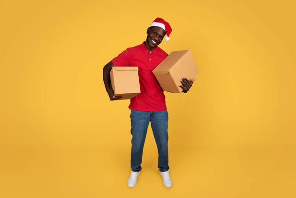 Счастливый молодой африканский курьер и Санта Клаус держат картонные коробки, изолированные на фоне оранжевой студии. Онлайн сервис доставки заказов, объявлений и предложений, работа на Рождество, Новый год - Фото, изображение