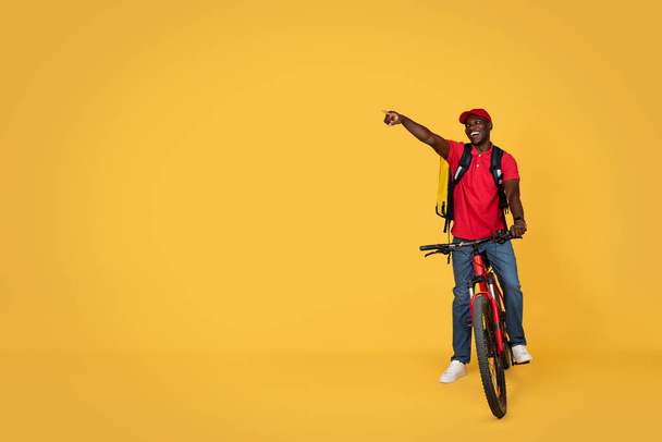 Ευτυχισμένος νεαρός Αφροαμερικάνος ντελιβεράς με στολή και σακίδιο με ποδήλατο, δείχνει τον ελεύθερο χώρο, απομονωμένος σε πορτοκαλί φόντο, ολόσωμος. Υπηρεσίες παράδοσης παραγγελιών, εργασίες σύστασης - Φωτογραφία, εικόνα