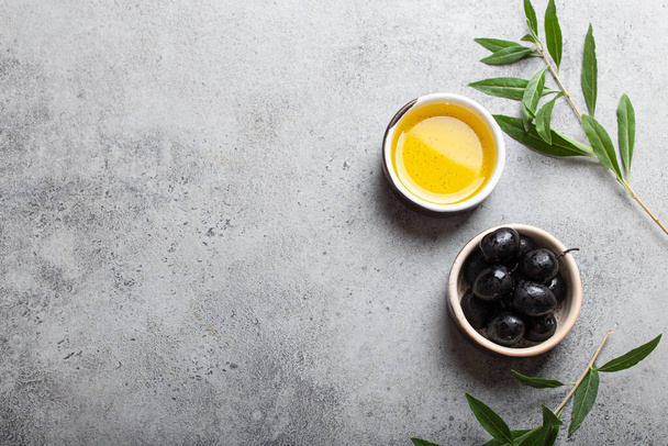 Black olives and extra virgin olive oil in little bowl with olive tree branches top view on gray concrete stone background, space for text - Photo, Image