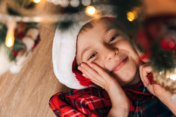 Un garçon joyeux en pyjama rouge-vert et un chapeau de Père Noël se trouve sur le sol. Noël à la maison. Vue du dessus - Photo, image