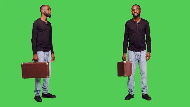 Случайный человек держит чемодан на камере и ждет чего-то, стоя на зеленом фоне экрана. Молодой человек с портфелем с багажом в студии, дружелюбный взрослый. - Фото, изображение