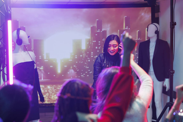 Люди на концерте техно-музыки в ночном клубе во время выступления диджея. Женщина-музыкант держит наушники во время смешивания звука с помощью контроллера во время вечеринки в ночном клубе - Фото, изображение