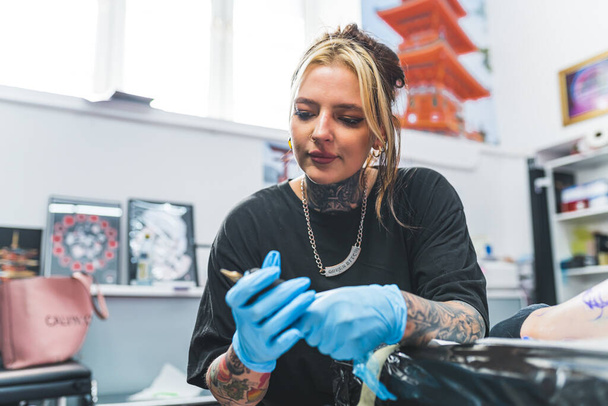 Επαγγελματική γυναίκα tattooist προετοιμασία του εξοπλισμού της και την αλλαγή μελάνι σε τατουάζ όπλο κατά τη διάρκεια ενός τατουάζ πόδι συνεδρία. Υψηλής ποιότητας φωτογραφία - Φωτογραφία, εικόνα