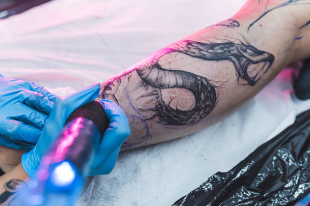 Creatura simile a un drago tatuata su una gamba di clienti caucasici da un tatuatore professionista con guanti di gomma protettivi. Foto di alta qualità - Foto, immagini