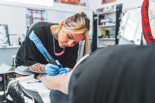 Взрослая профессиональная женщина-татуировщица татуирует ноги молодых женщин. Сосредоточенная художница с татуировочным пистолетом. Высокое качество фото - Фото, изображение