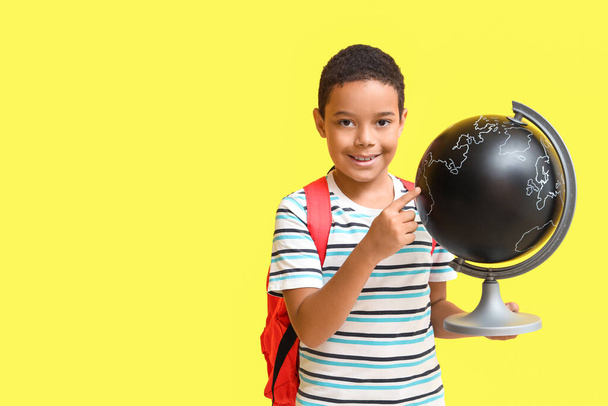 Μικρό Αφροαμερικανό αγόρι με σχολική τσάντα που δείχνει την υδρόγειο σε κίτρινο φόντο - Φωτογραφία, εικόνα