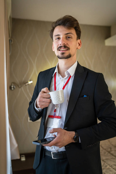 Jeden mężczyzna dorosły biały mężczyzna w garniturze z wąsami i brodą biznesmen w pokoju hotelowym trzymać filiżankę kawy przed lub po pracy biorąc hamulec prawdziwi ludzie kopiować przestrzeń - Zdjęcie, obraz