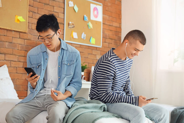 Studenti di sesso maschile con auricolari utilizzando i telefoni cellulari in camera dormitorio - Foto, immagini