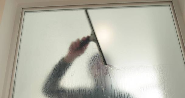 Ένας άντρας ακουμπάει μια παγωμένη επίστρωση σε μια γυάλινη πόρτα σε μια κλινική για να κάνει την γυάλινη πόρτα αδιαφανή. Εφαρμογή ματ επίστρωσης στο γυαλί. - Φωτογραφία, εικόνα