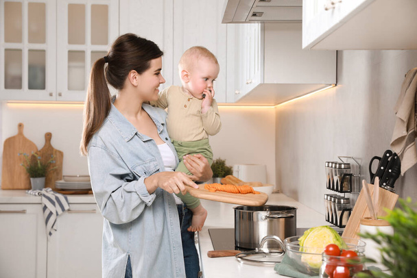 Happy young woman holding her cute little baby while cooking in kitchen - Фото, зображення