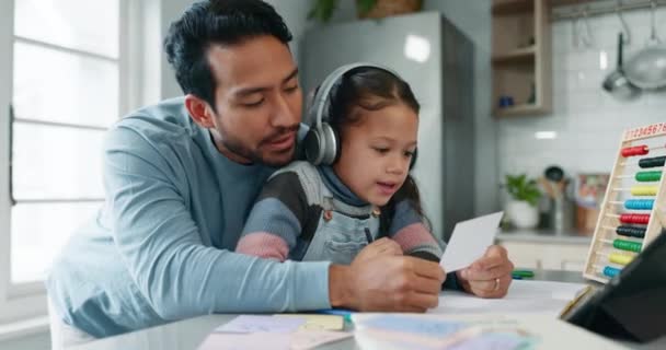 Home school help, father and child with reading card, headphones and tablet, learning app and education in house. Elearning, studying and dad teaching girl with homework in kitchen for online class - Footage, Video