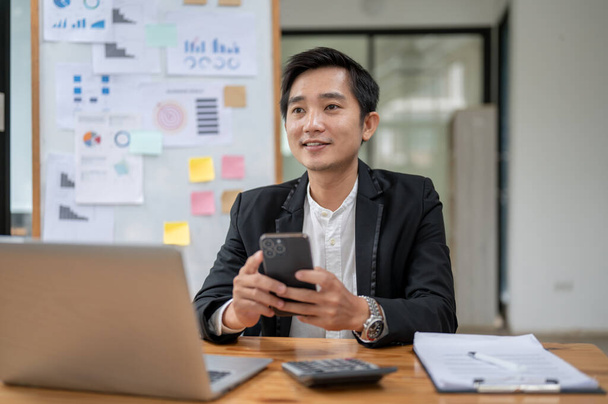 Szczęśliwy i profesjonalny biznesmen azjatycki za pomocą smartfona przy biurku w biurze. - Zdjęcie, obraz