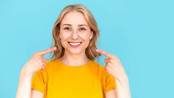 Szczęśliwa kobieta wskaż palcem wskazującym biały uśmiech, zademonstruj jej zdrowe i doskonałe zęby, stojąc odizolowane na niebieskim tle przestrzeni kopii. Koncepcja leczenia stomatologicznego, pielęgnacji jamy ustnej, pielęgnacji skóry, baner internetowy - Zdjęcie, obraz